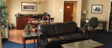 A living room with a leather sofa, a glass-top coffee table, a dining area with a wooden table and chairs, and a kitchen in the background.