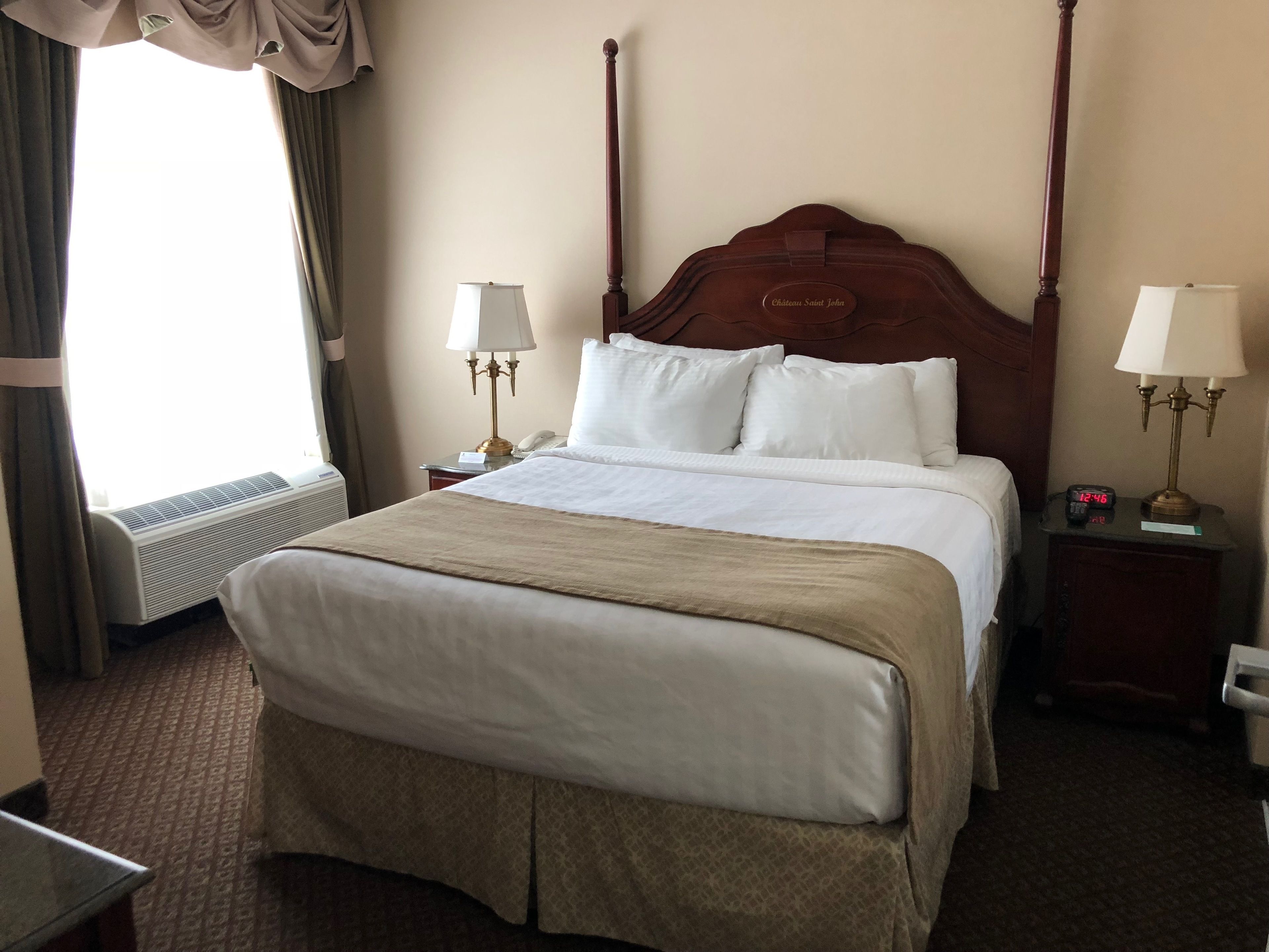A hotel room with a large bed, two bedside tables with lamps, a bedside clock, a window with curtains, and an air conditioning unit.