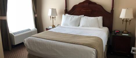 A hotel room with a large bed, two bedside tables with lamps, a bedside clock, a window with curtains, and an air conditioning unit.