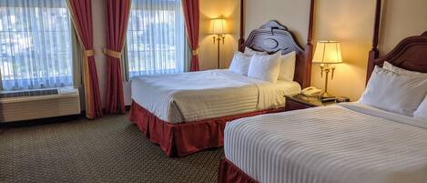 A hotel room with two beds, a large window with curtains, a bedside table with a lamp, and a wall-mounted telephone.