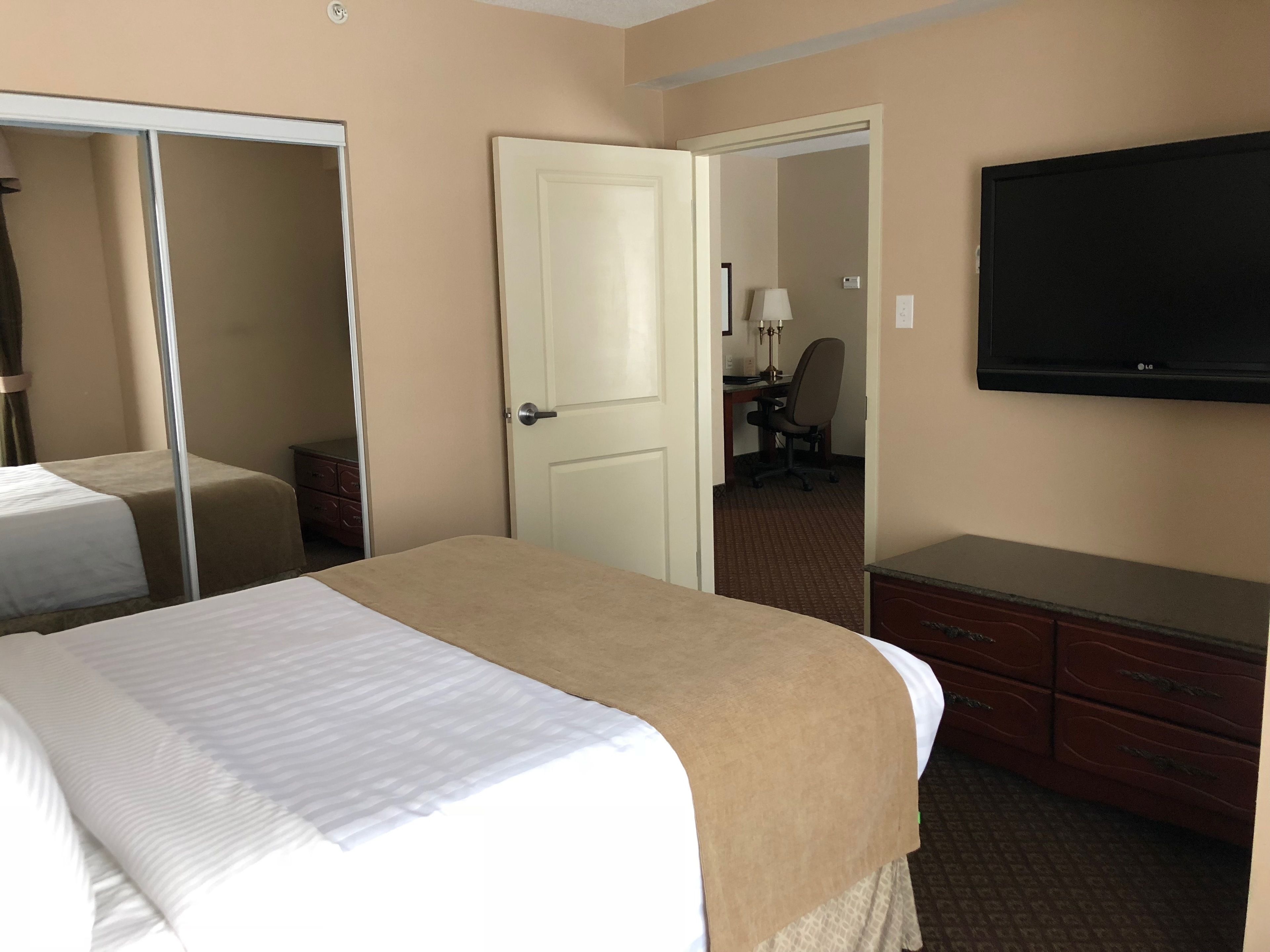 A hotel room with a bed, a dresser, a TV, and a door leading to another room.