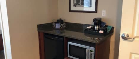 Una kitchenette compacta con microondas, cafetera y copas de vino sobre la encimera.
