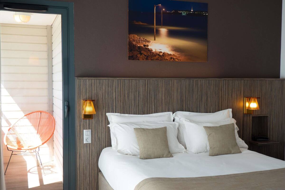A hotel room with a bed, two wall-mounted lamps, a night view painting, and a chair by the door.