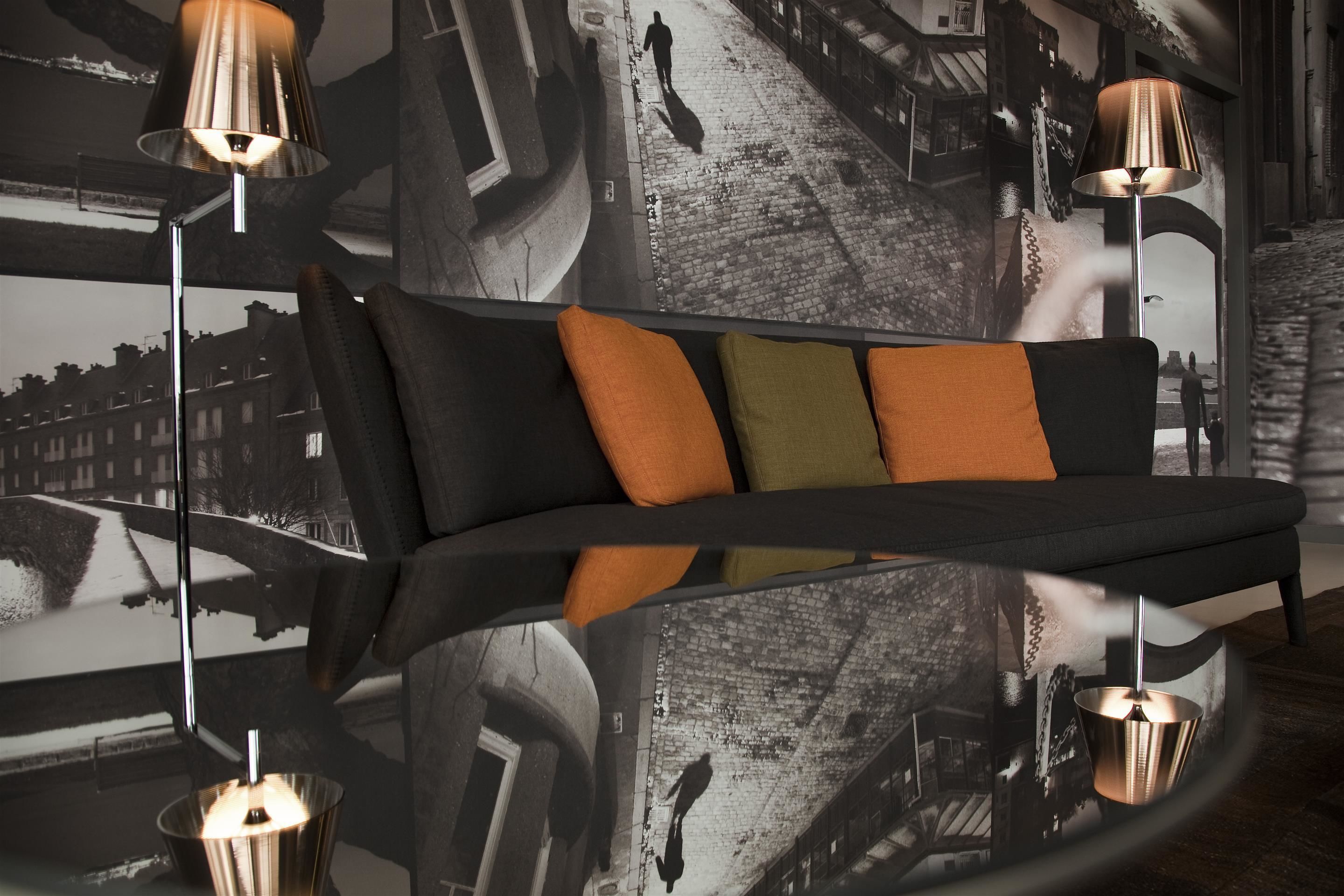 A modern room with a reflective glass table, a black sofa with orange and green cushions, and two lamps with metallic shades.
