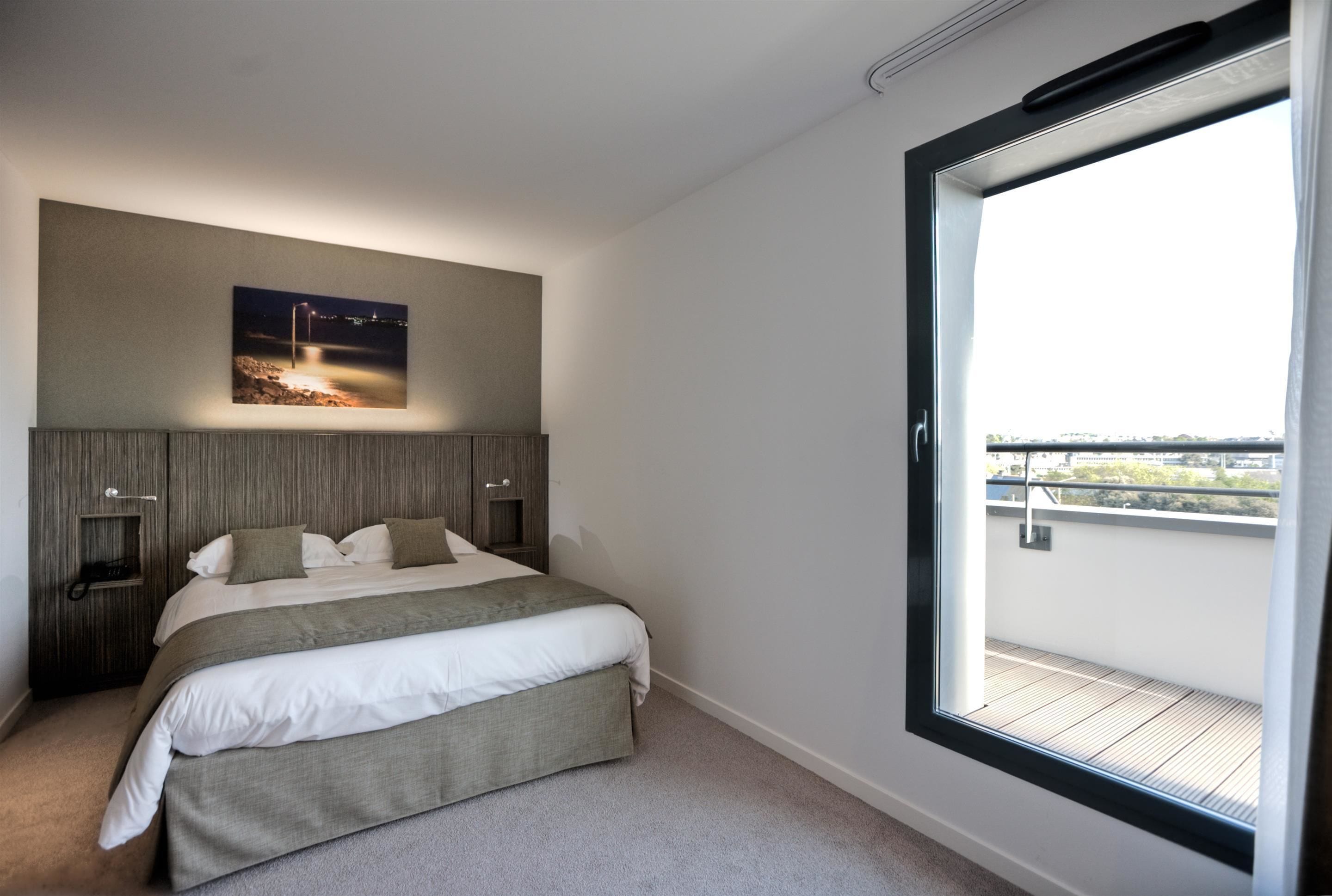 Un dormitorio moderno con una cama grande, una mesita de noche, un estante en la pared y un amplio ventanal con vistas a la ciudad.