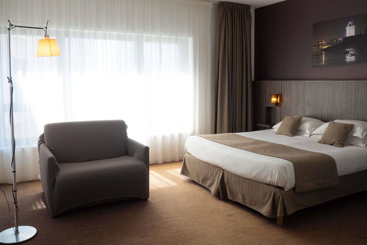 A hotel room with a bed, a chair, a floor lamp, and a painting on the wall.