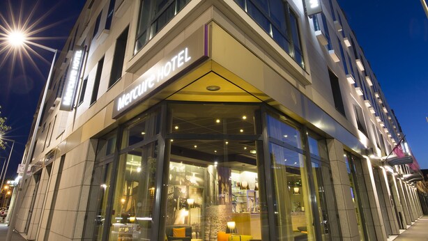 Un hôtel moderne doté d’une façade en verre, d’un hall d’entrée spacieux avec des sièges et d’une enseigne indiquant « Hôtel Mercure ».