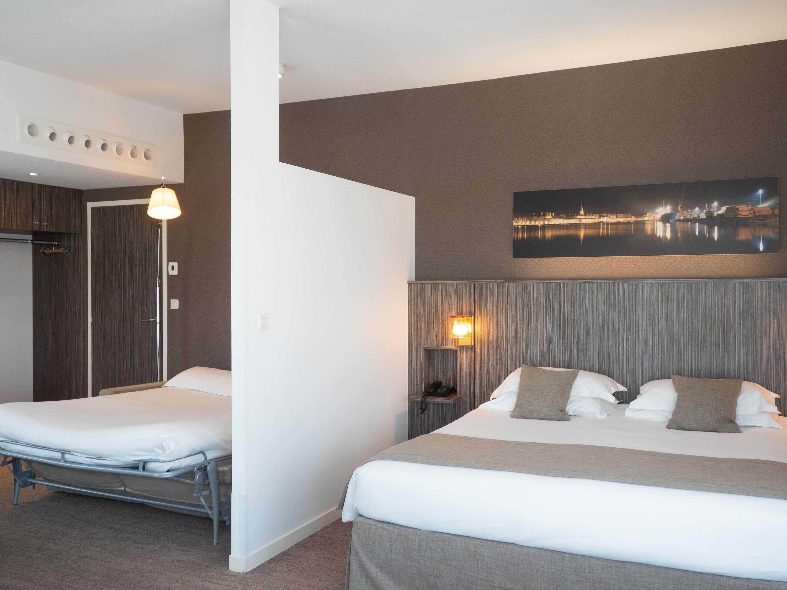 A hotel room with a bed, bedside table, lamp, and a cityscape painting on the wall.