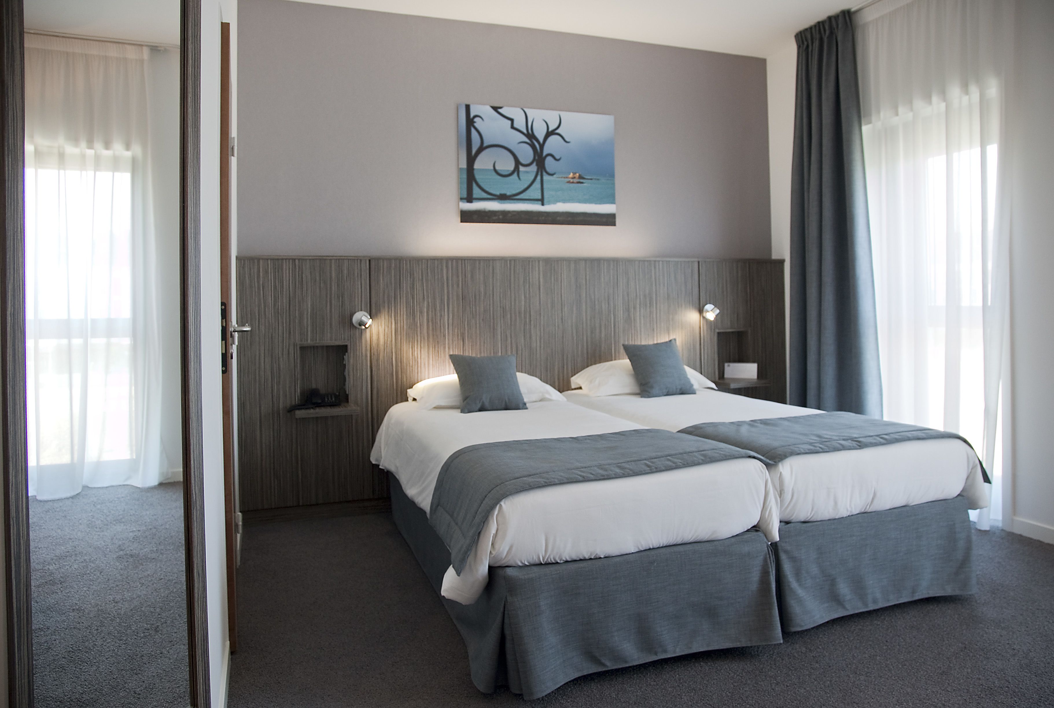 A modern hotel room with two beds, a large artwork on the wall, and a view of the sea through the window.