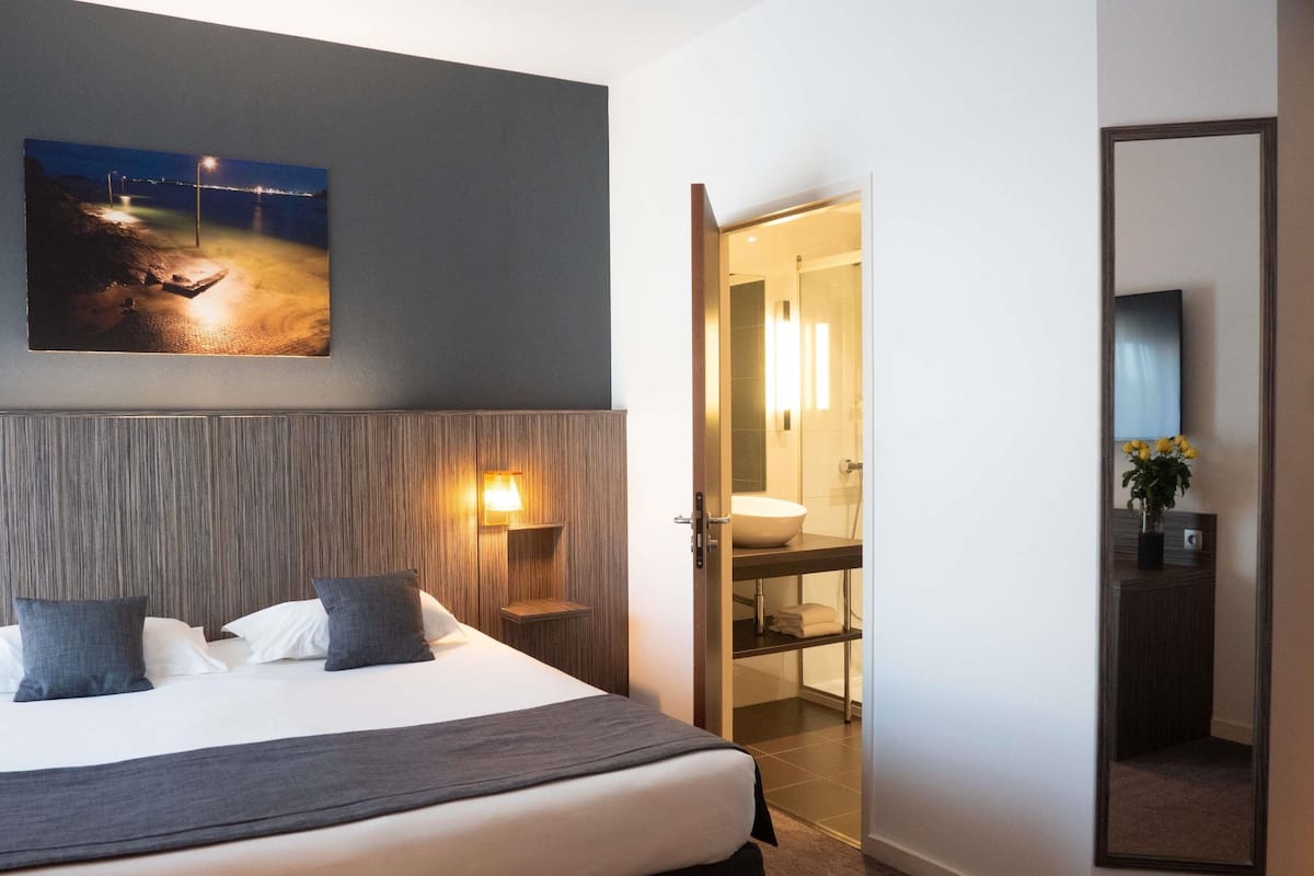 A hotel room with a bed, a night view painting, a nightstand with a lamp, a bathroom with a sink and mirror, and a large mirror.