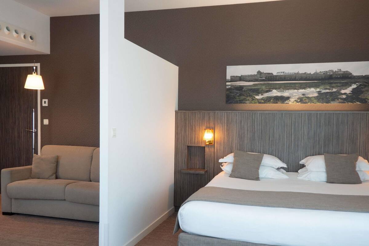 A hotel room with a bed, a sofa, a nightstand with a lamp, a painting on the wall, and a door.