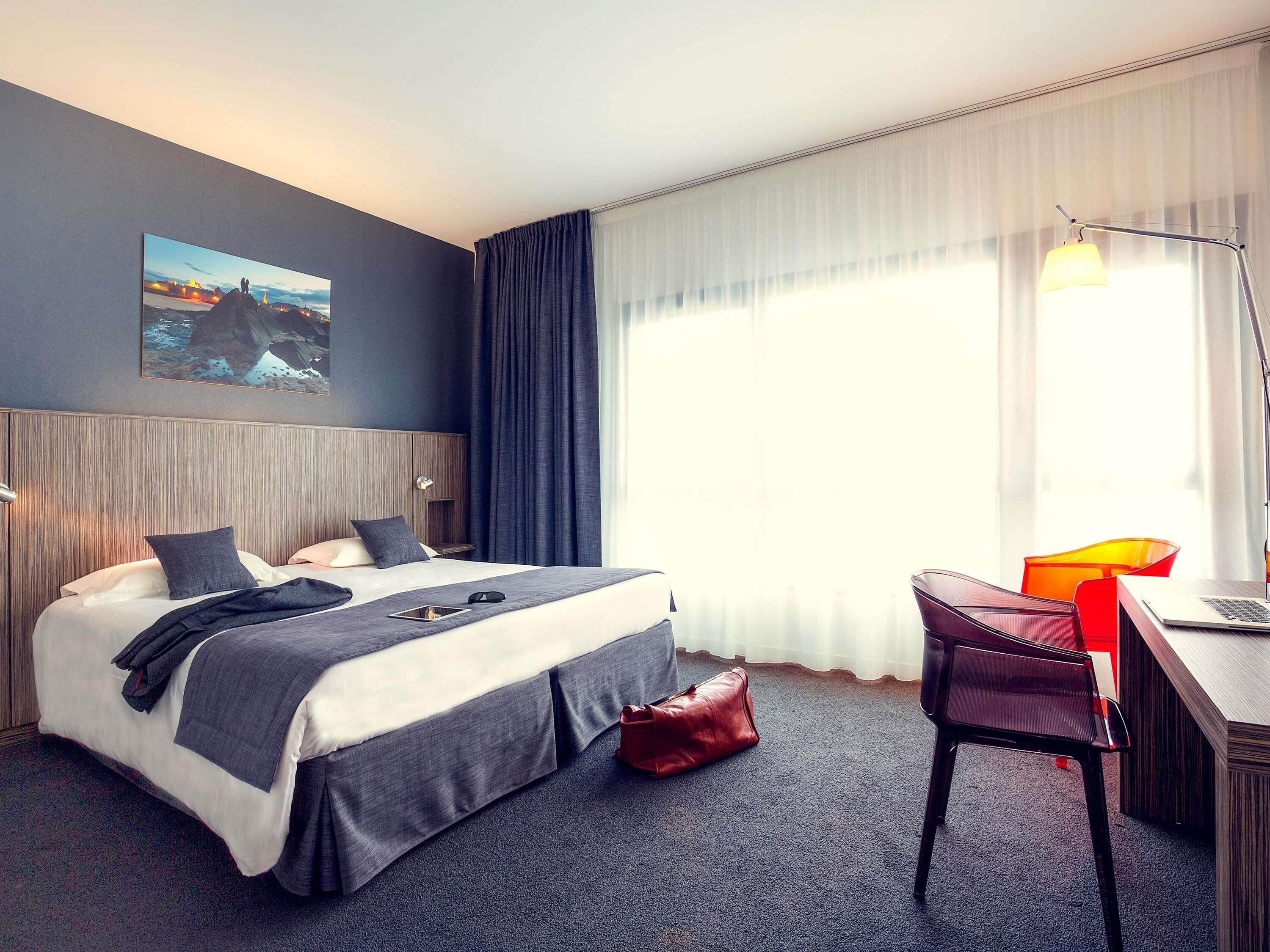 A hotel room with a bed, a desk, a chair, a lamp, and a painting on the wall.