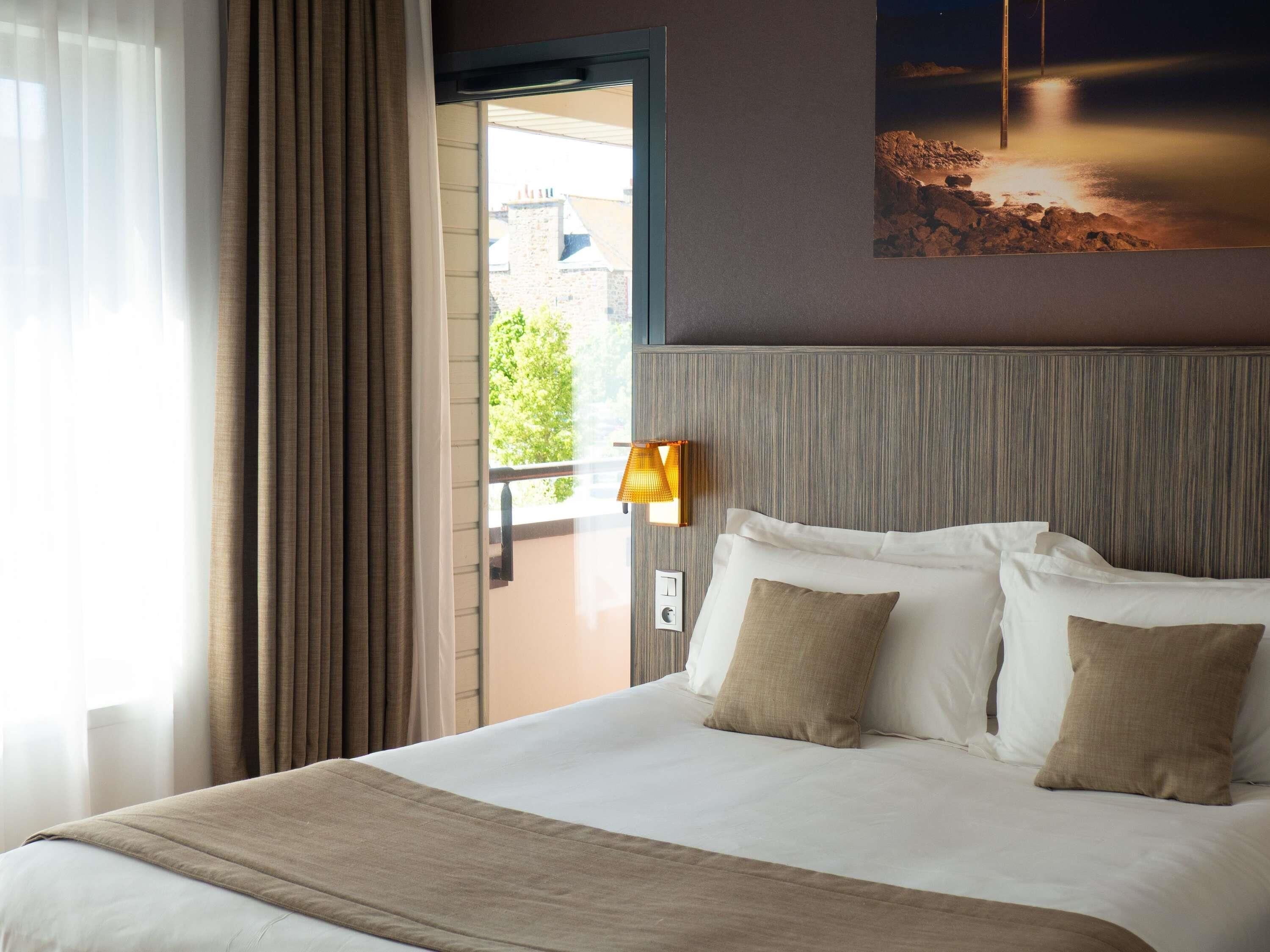 Quarto de hotel com uma cama, cortinas, janela e uma luminária fixada na parede.