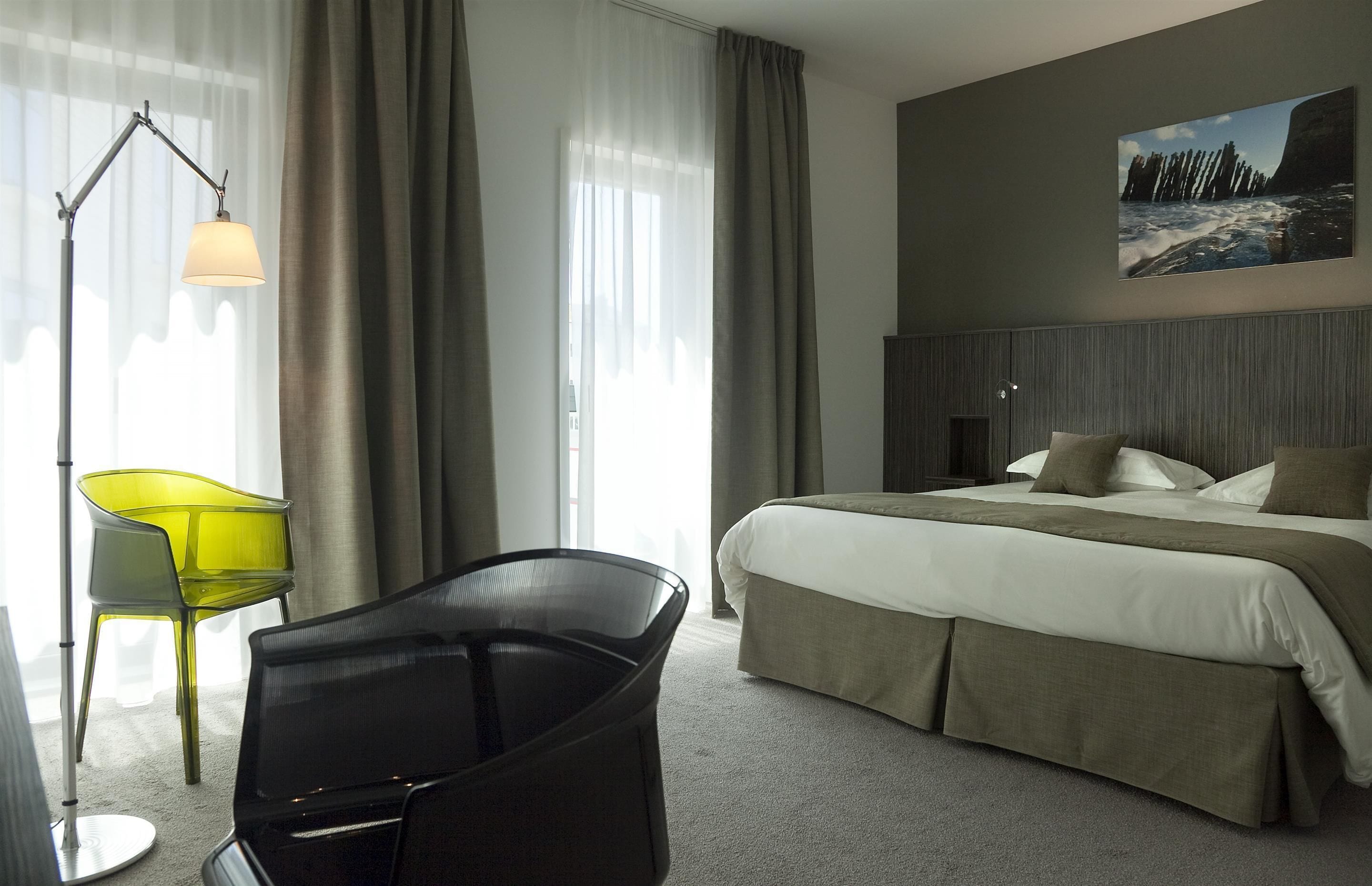 Un cuarto de hotel con cama, escritorio, silla, lámpara y un cuadro en la pared.