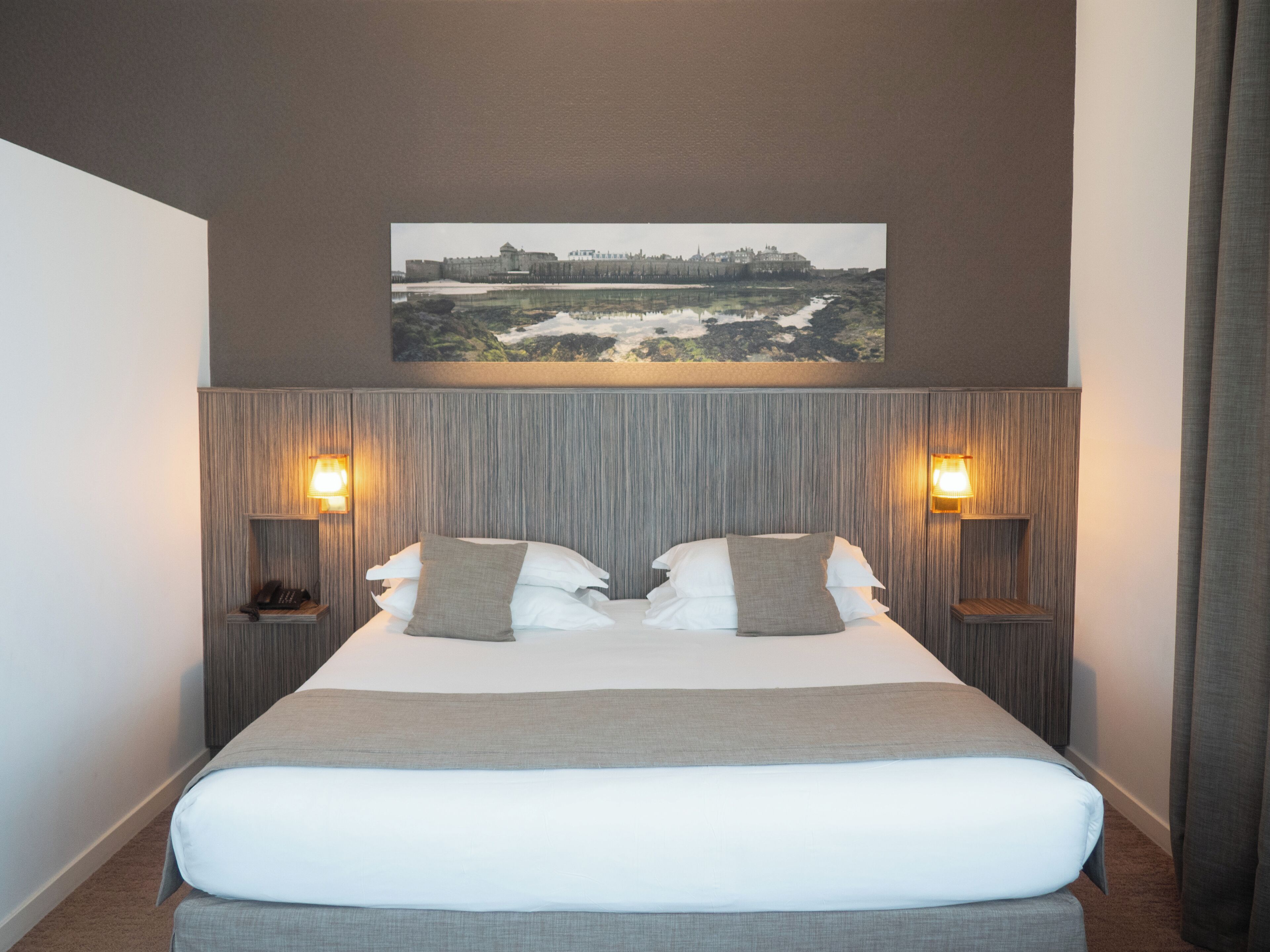 A hotel room with a large bed, two bedside tables, a nightstand, a wall-mounted lamp, and a framed picture above the bed.