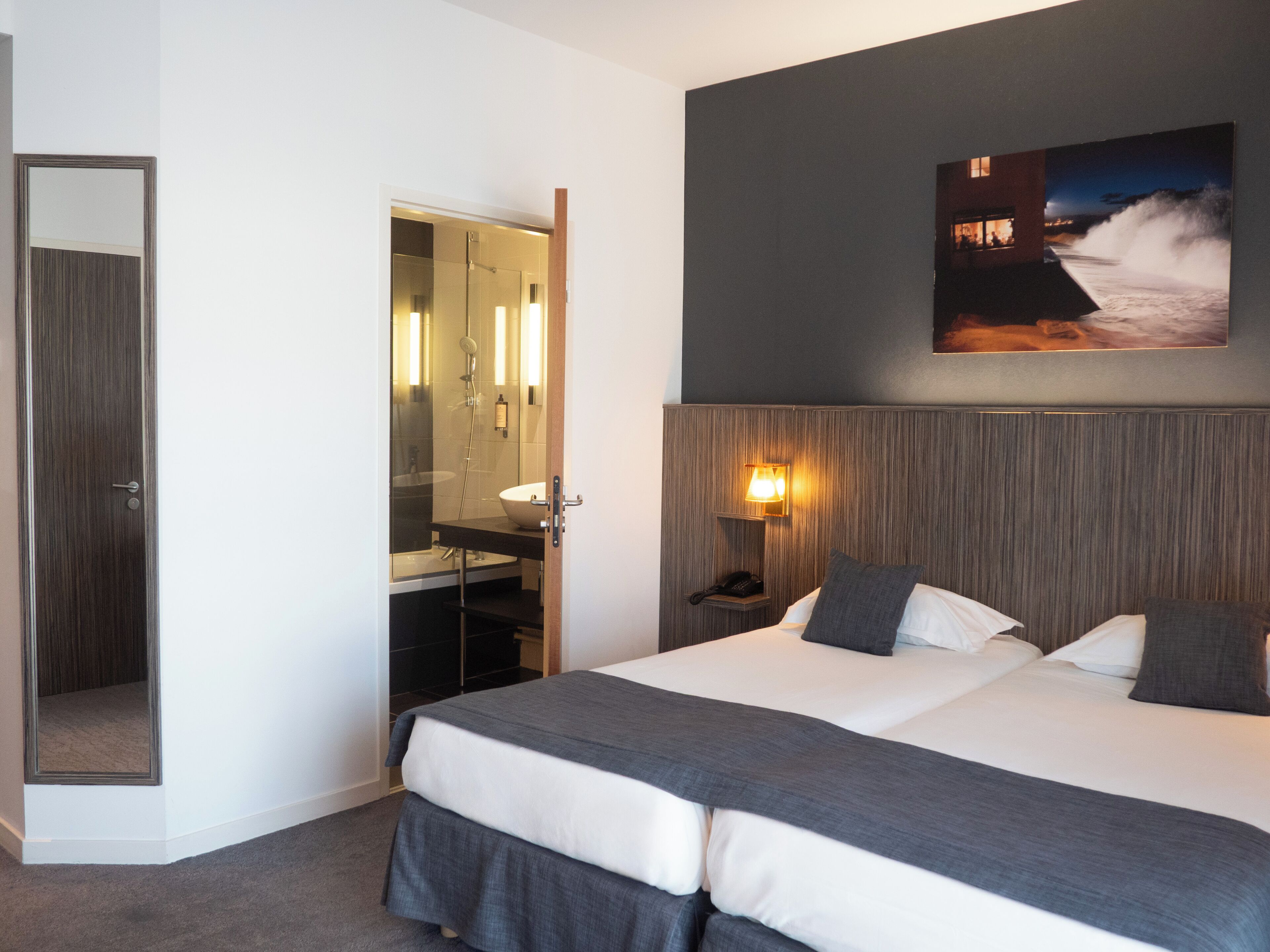 A hotel room with a bed, a nightstand, a lamp, a mirror, and a painting on the wall.