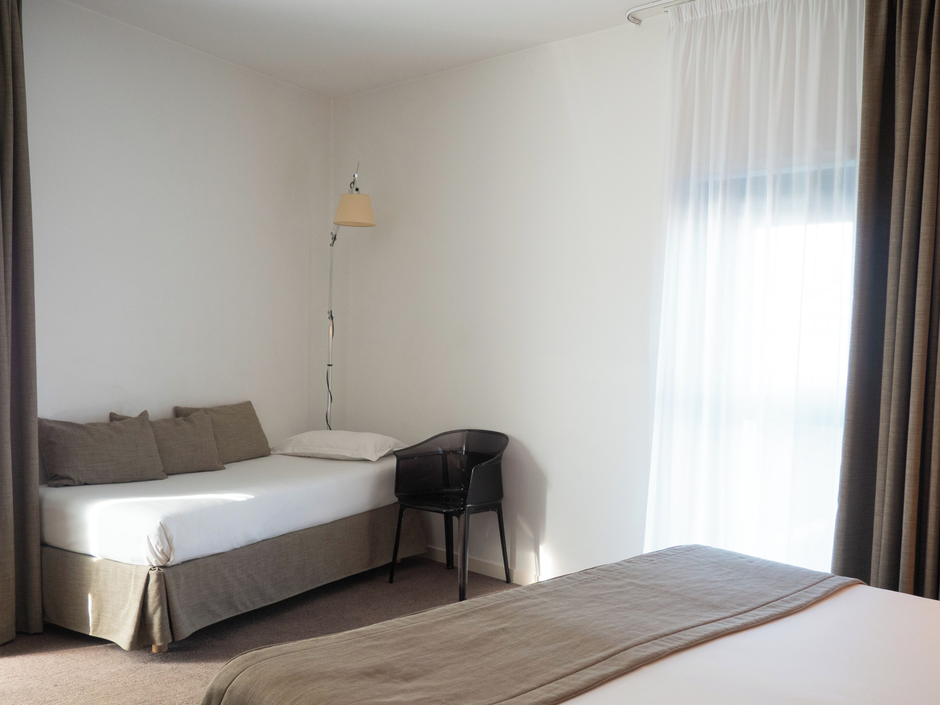 A single bed with a headboard, a bedside table, a lamp, and a chair in a room with a window and curtains.