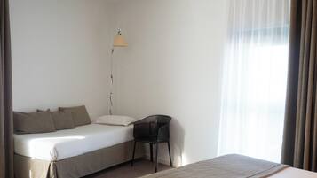 A single bed with a headboard, a bedside table, a lamp, and a chair in a room with a window and curtains.