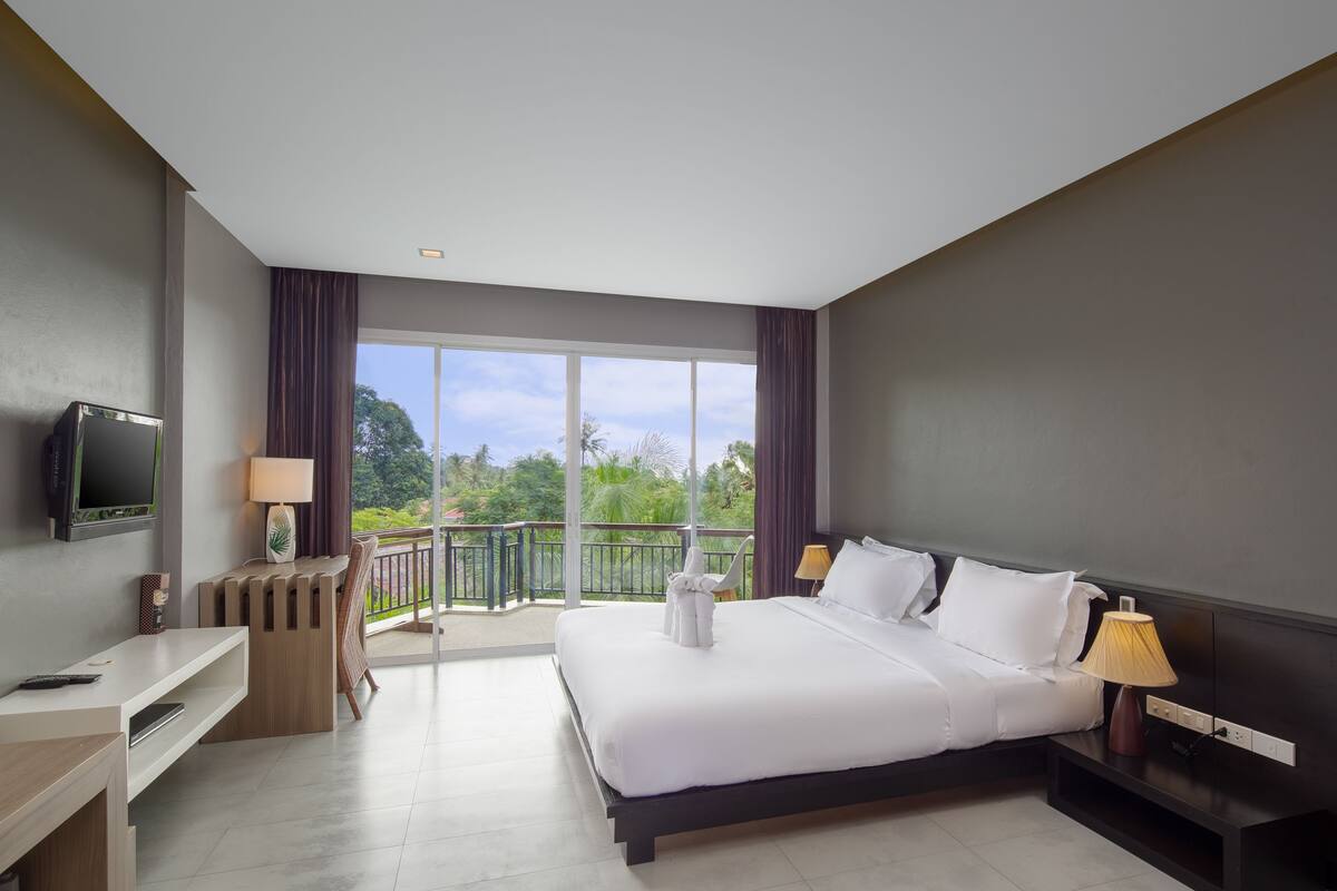 Una habitación de hotel con una cama grande, un escritorio, un televisor y un balcón con vistas a zonas verdes.