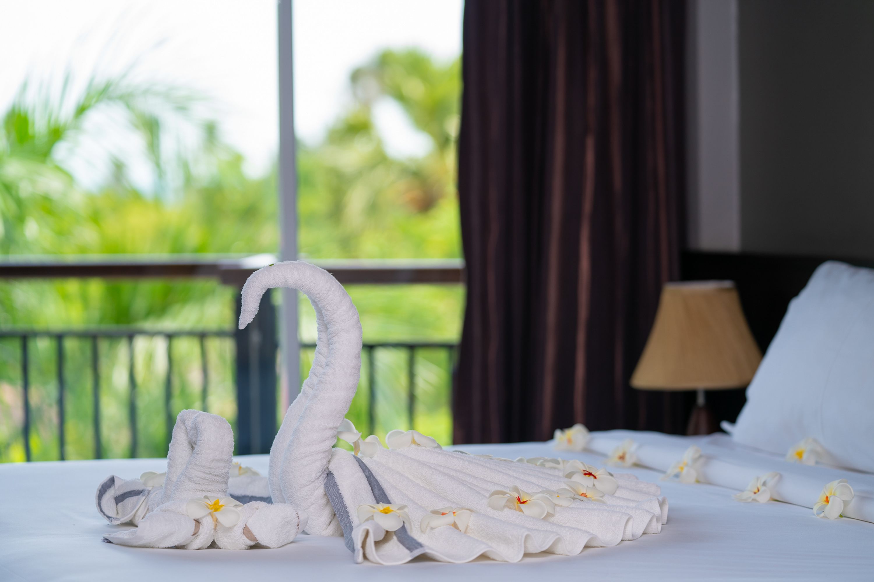 Un lit parfaitement fait, agrémenté d’une serviette pliée en forme de cygne et de fleurs blanches, le tout mis en valeur par un balcon verdoyant en arrière-plan.