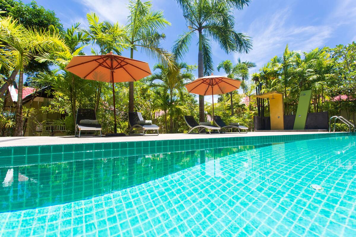 Una piscina con bordes de azulejos, rodeada de sillas de descanso y sombrillas, con palmeras y un cielo despejado al fondo.