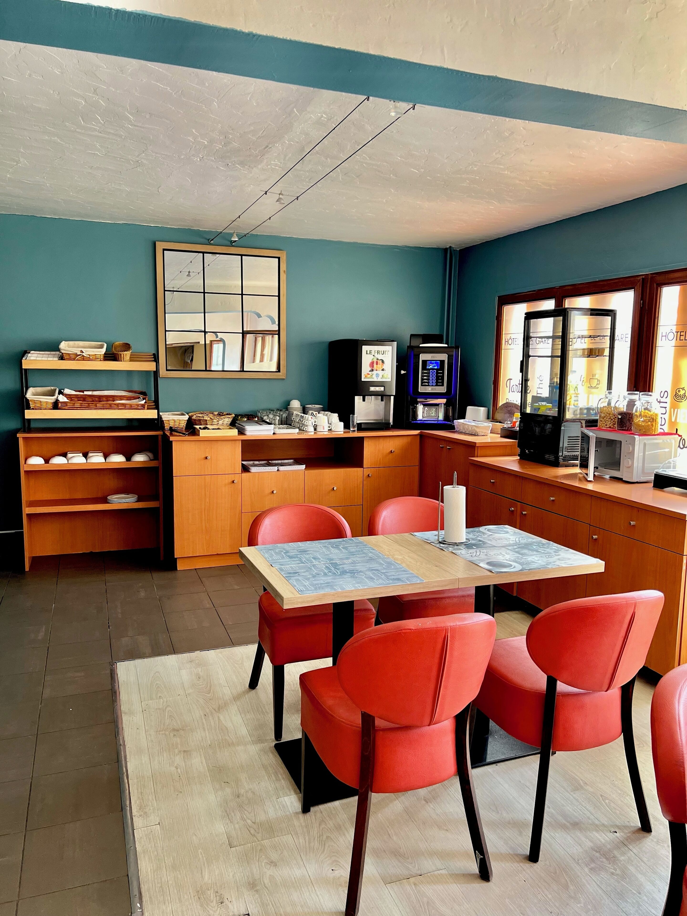 A compact hotel room with a wooden table and red chairs, a kitchenette with a coffee machine, and a large window with a view of the hotel.