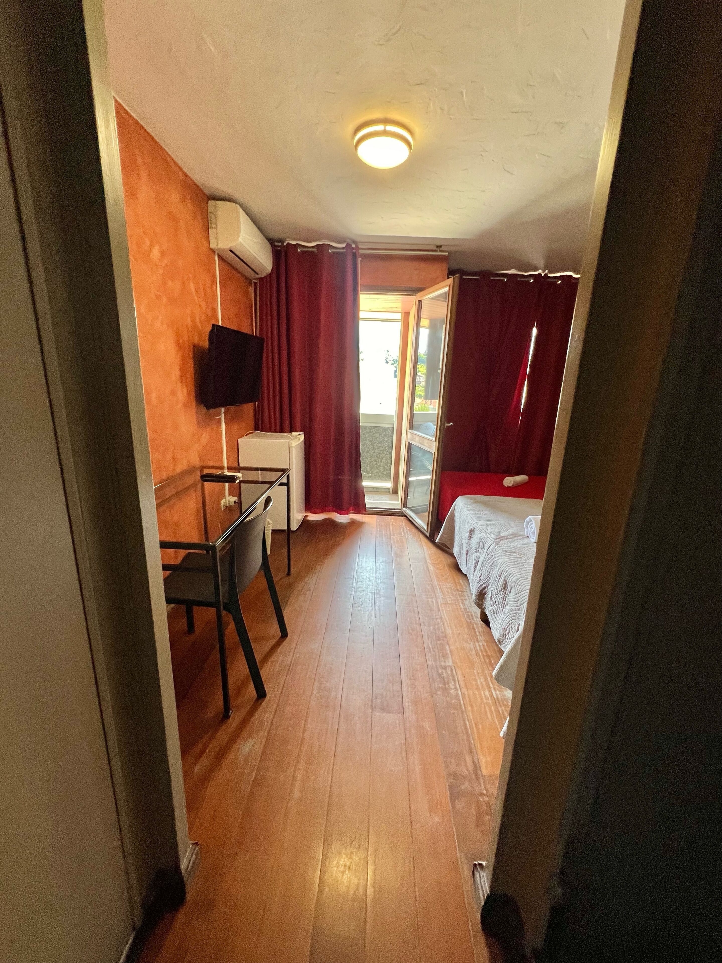 Une chambre d’hôtel de taille modeste, avec un plancher en bois, un bureau avec une chaise, un lit avec des rideaux rouges et un téléviseur fixé au mur.