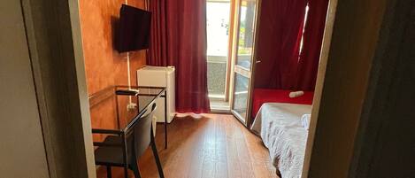 Une chambre d’hôtel de taille modeste, avec un plancher en bois, un bureau avec une chaise, un lit avec des rideaux rouges et un téléviseur fixé au mur.