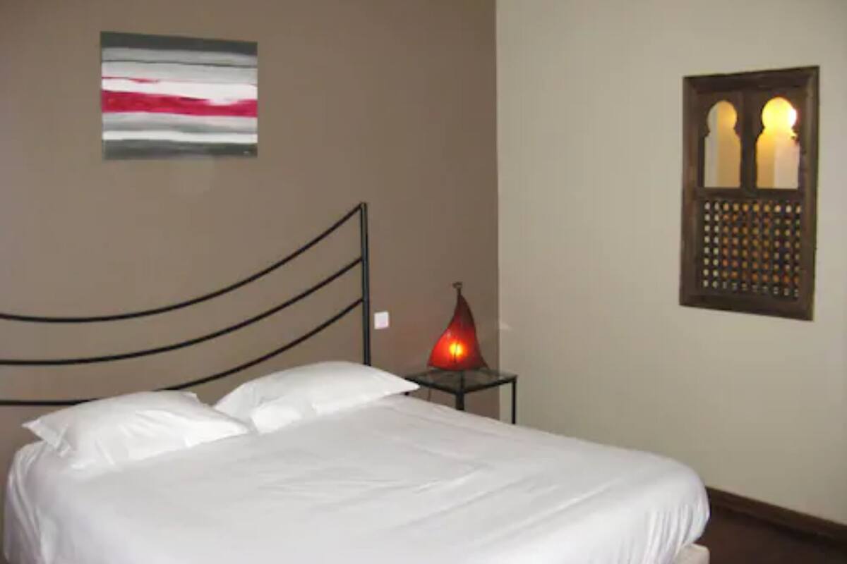 A bedroom with a bed, a wall-mounted artwork, a window with a decorative grille, and a bedside table with a lamp.