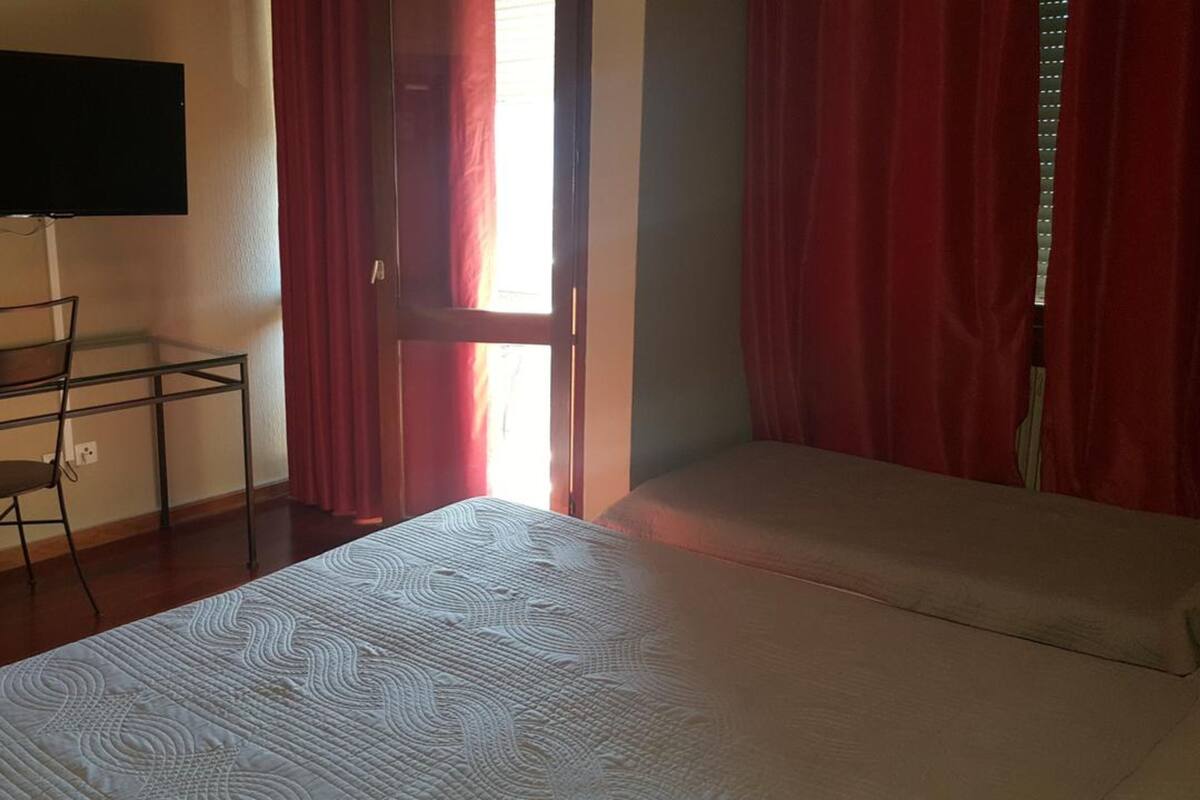 A hotel room with a bed, a desk with a chair, a TV mounted on the wall, and red curtains.