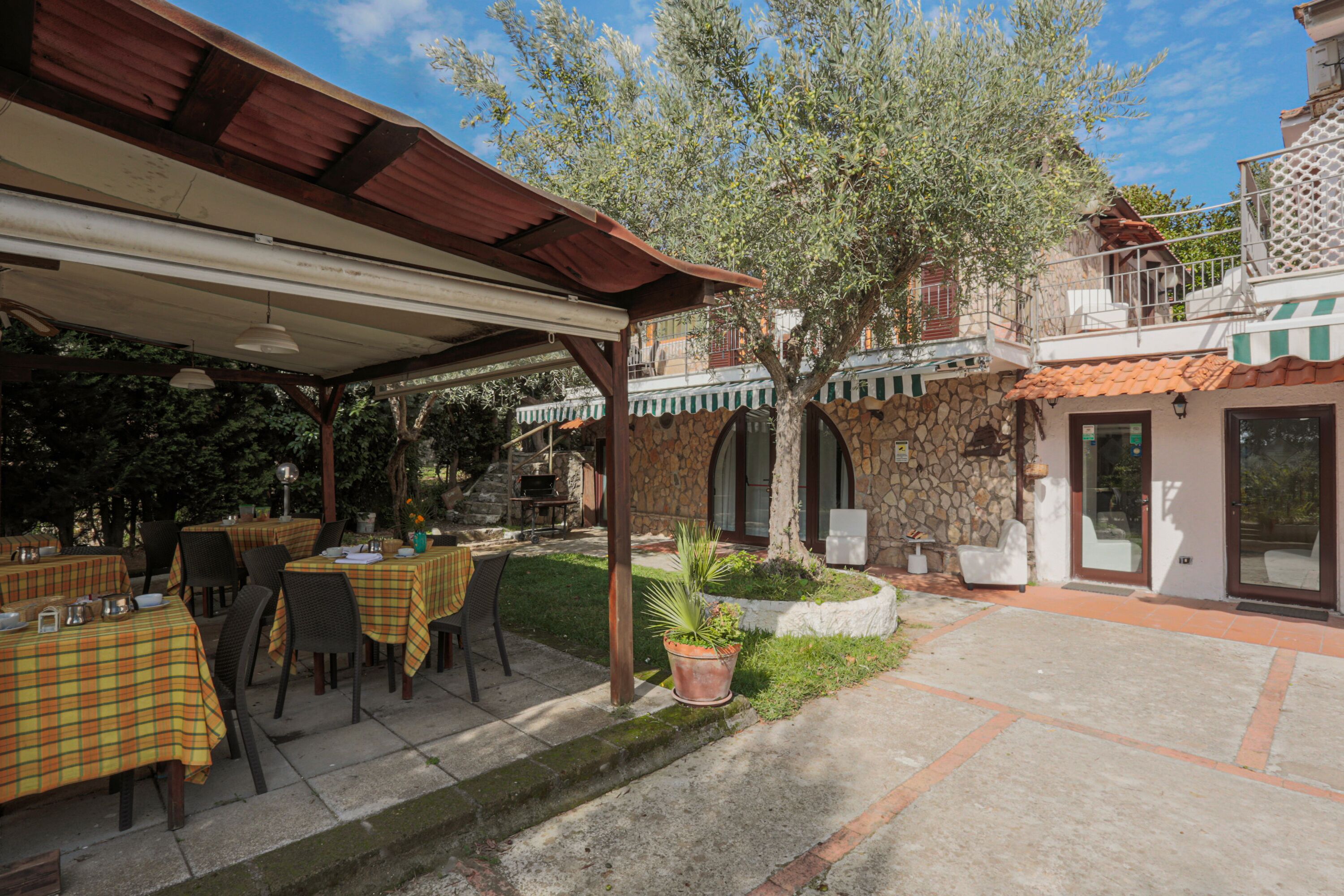 Un área de comedor al aire libre con mesas y sillas, un espacio de estar cubierto y un edificio de piedra con balcón.
