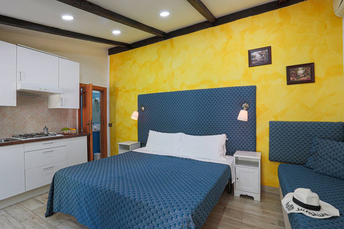 A bedroom with a bed, bedside tables, a nightstand, a lamp, and a kitchen area with cabinets and a stove.