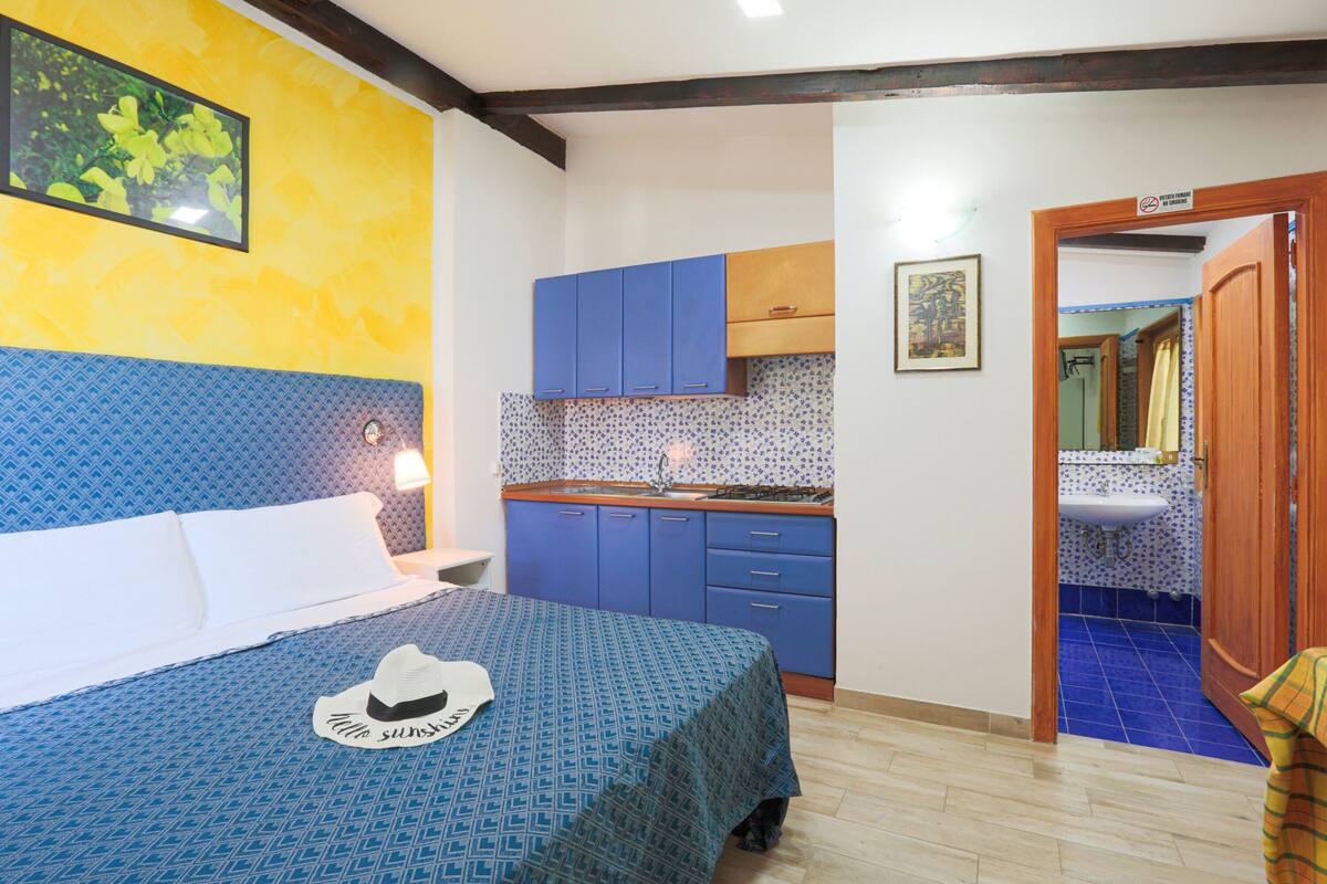 A hotel room with a bed, a blue patterned bedspread, a hat on the bed, a kitchenette with blue cabinets, and a bathroom visible through an open door.