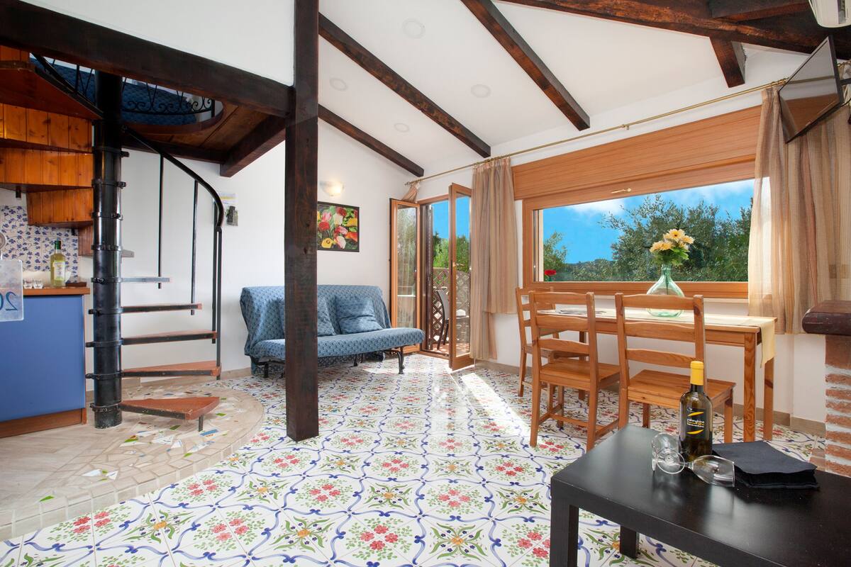 A room with a wooden dining table, chairs, a sofa, and a kitchen area with a stove.