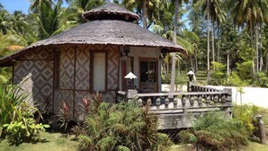 Une cabane à toit de chaume, dotée d’un porche en bois, entourée d’une végétation tropicale et de palmiers.