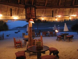 Un espacio al aire libre con mesas y sillas, techo de paja y vista a la playa y al mar.
