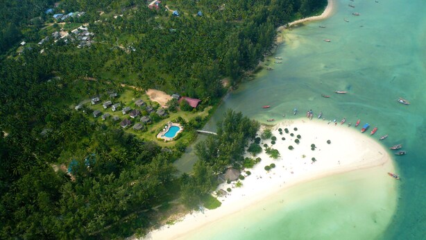 Vue aérienne d’un complexe hôtelier tropical doté d’une vaste plage de sable, d’une piscine et de plusieurs bateaux sur l’eau.