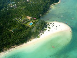 Vista aérea de un resort tropical con una amplia playa de arena, una piscina y varias embarcaciones en el agua.