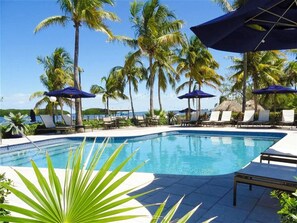 Un área junto a la piscina con sillones de descanso y sombrillas, rodeada de palmeras.