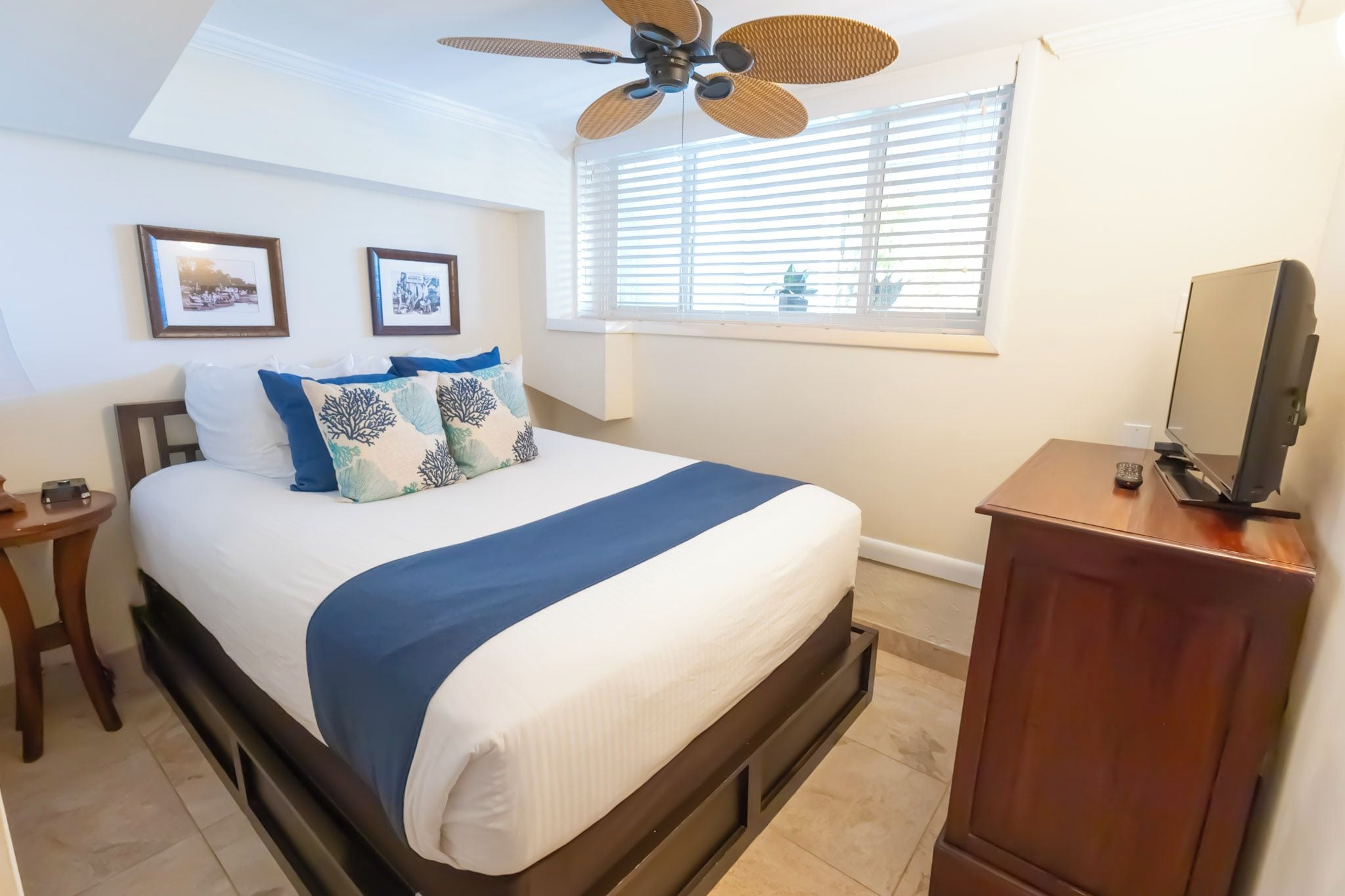 Un dormitorio con una cama, ventilador de techo, ventana con persianas, televisor y cómoda de madera.