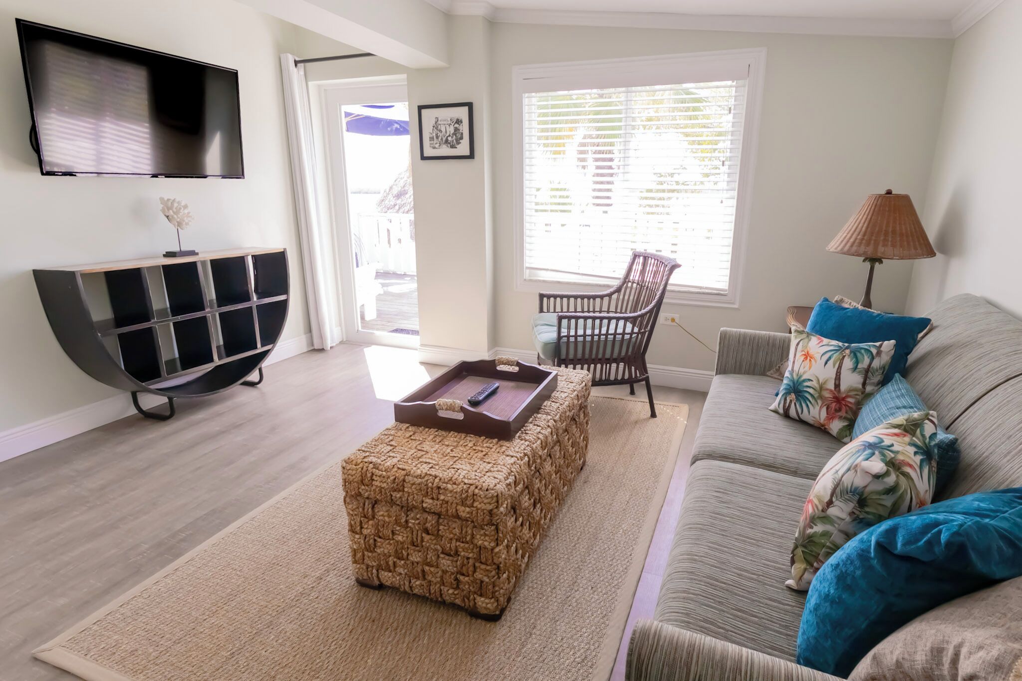 A living room with a flat-screen TV, a woven ottoman, a sofa with floral cushions, a wicker chair, and a lamp.