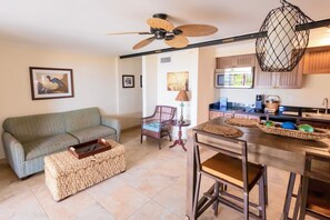 Sala de estar con sofá, mesa de centro, reposapiés de mimbre, una silla, mesa de comedor y área de cocina con microondas y lámpara colgante.