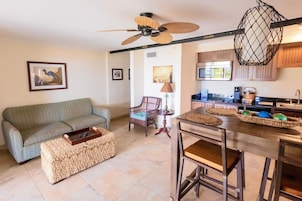 A living room with a sofa, a coffee table, a wicker ottoman, a chair, a dining table, and a kitchen area with a microwave and a hanging light fixture.