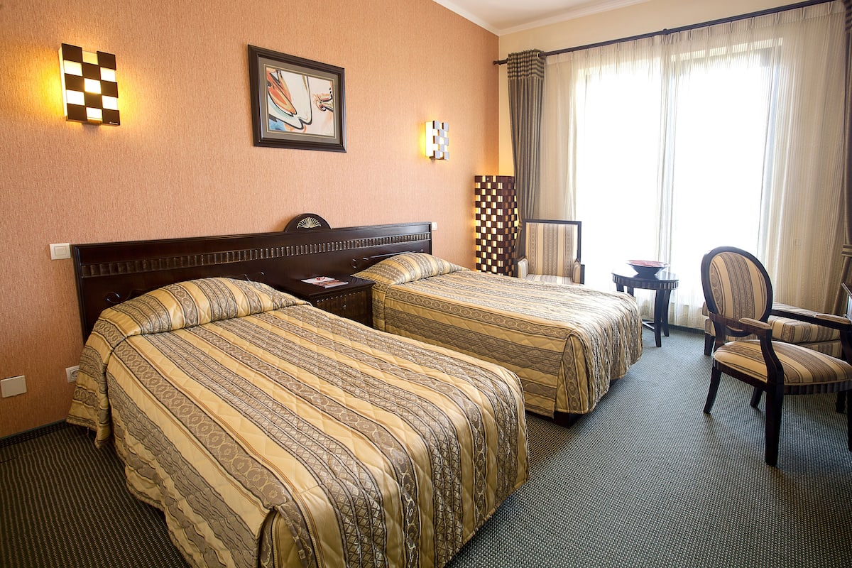 Ein Hotelzimmer mit zwei Betten, einem Schreibtisch, einem Stuhl und einem großen Fenster mit Vorhängen.