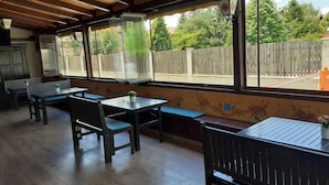 Un patio cubierto con mesas y bancos de madera, ventanas grandes con marcos de madera y vistas a un patio trasero cercado.