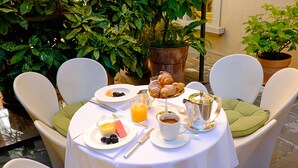 A table set for breakfast with croissants, fruit, and coffee, surrounded by white chairs with green cushions.