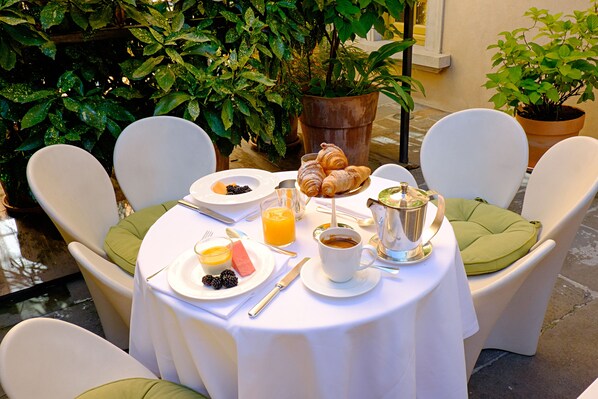 Une table dressée pour le petit-déjeuner, avec des croissants, des fruits et du café, entourée de chaises blanches dotées de coussins verts.