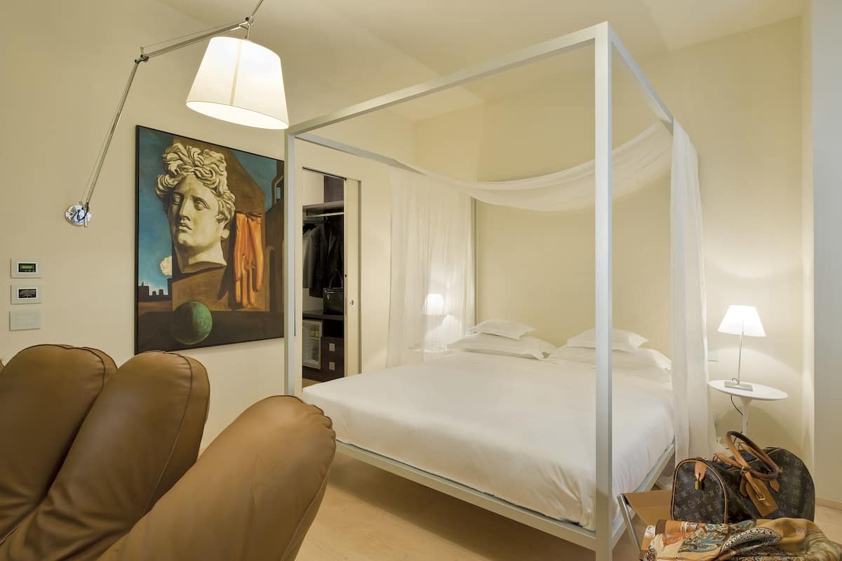 A four-poster bed with a canopy, a painting on the wall, a brown leather chair, and a bedside table with a lamp.