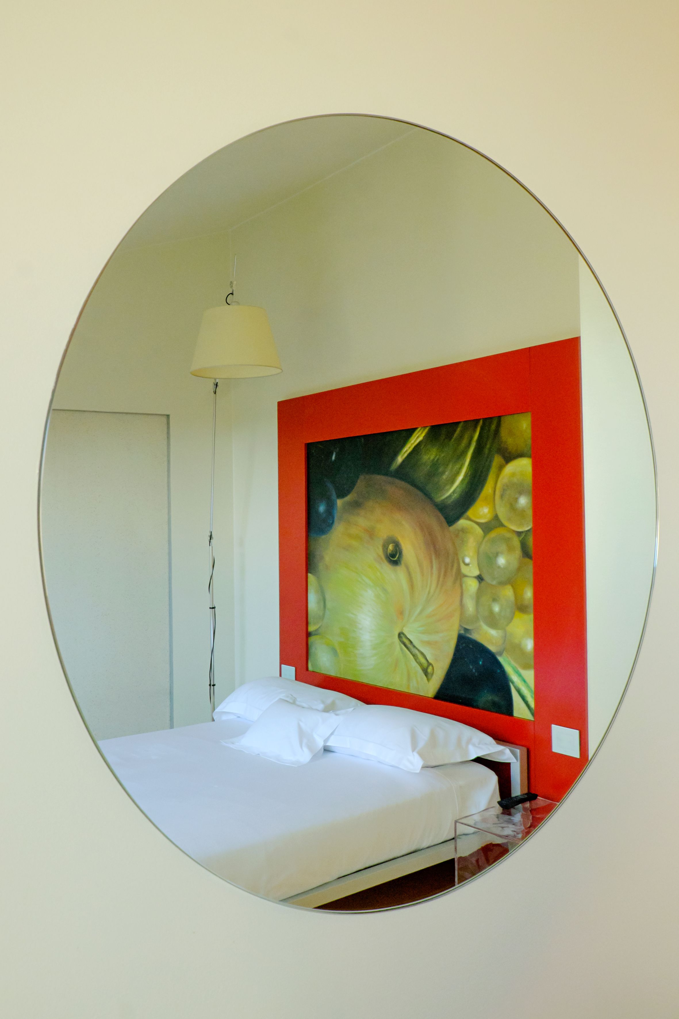 A circular mirror reflecting a bed with white bedding and a red-framed painting of fruits.
