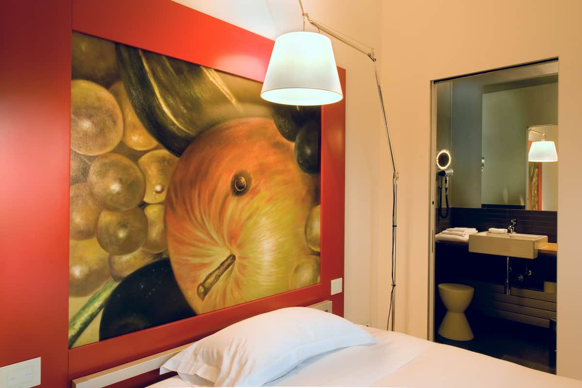 A hotel room with a large fruit-themed painting on the wall, a bed with white bedding, a modern bathroom with a mirror and sink.